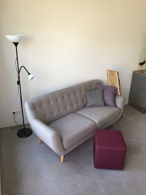 Un canapé gris aux pieds en bois, un pouf rouge, une lampe sur pied et une tête de lit en bois, le tout placé devant un mur uni.