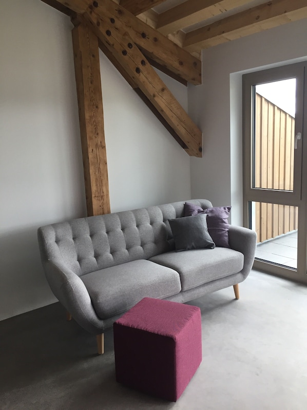 A grey sofa with cushions, a wooden beam ceiling, and a large window.