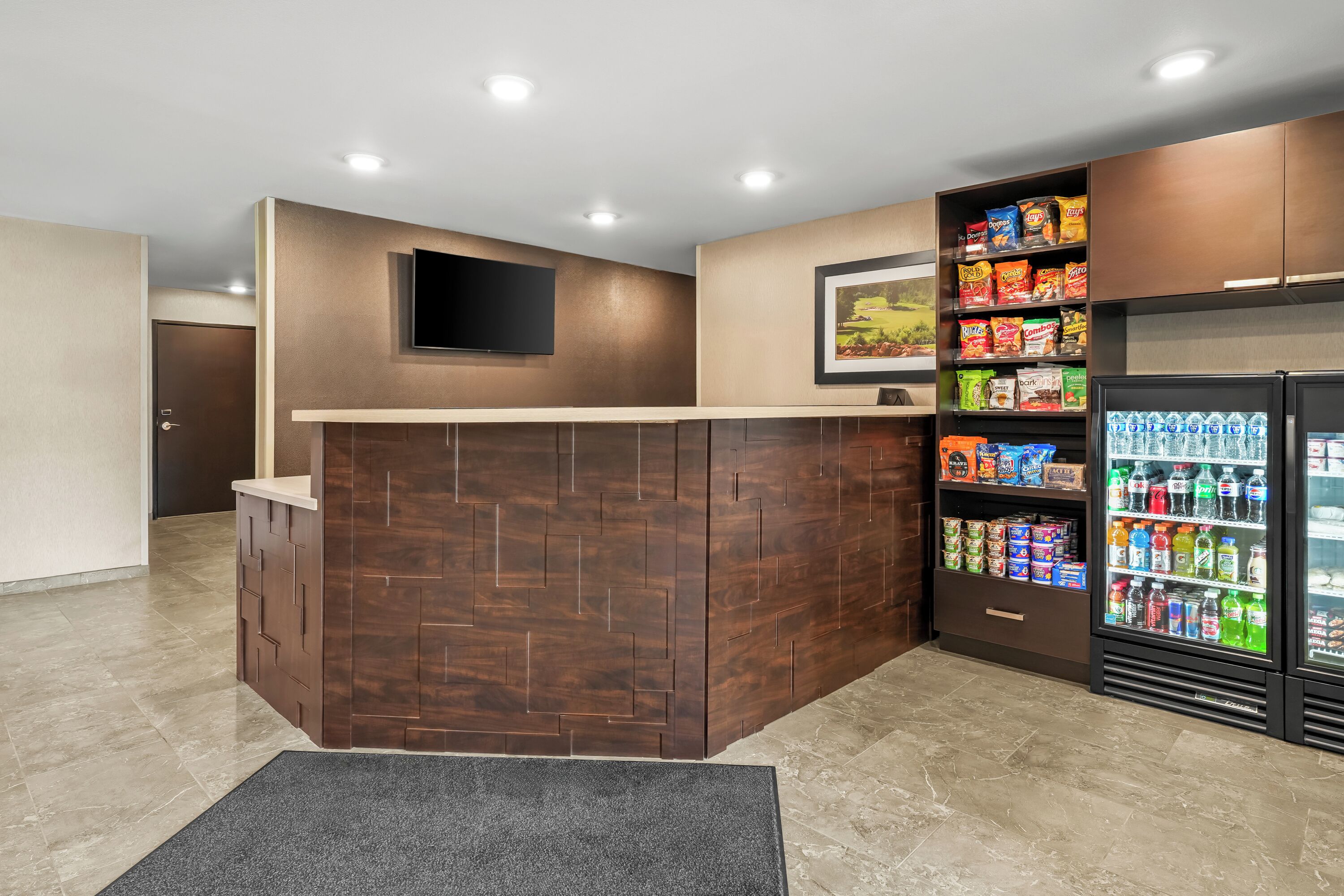 Un área de recepción con un mostrador de madera, un televisor de pantalla plana montado encima, un cuadro en la pared y una máquina expendedora llena de aperitivos y bebidas.