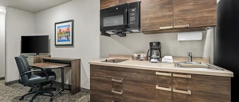A compact kitchen with wooden cabinets, a microwave, a coffee maker, and a small dining area with a chair and table.