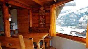 Interior de una cabaña de madera con una mesa y sillas, una ventana con cortinas y vistas a una casa de piedra.