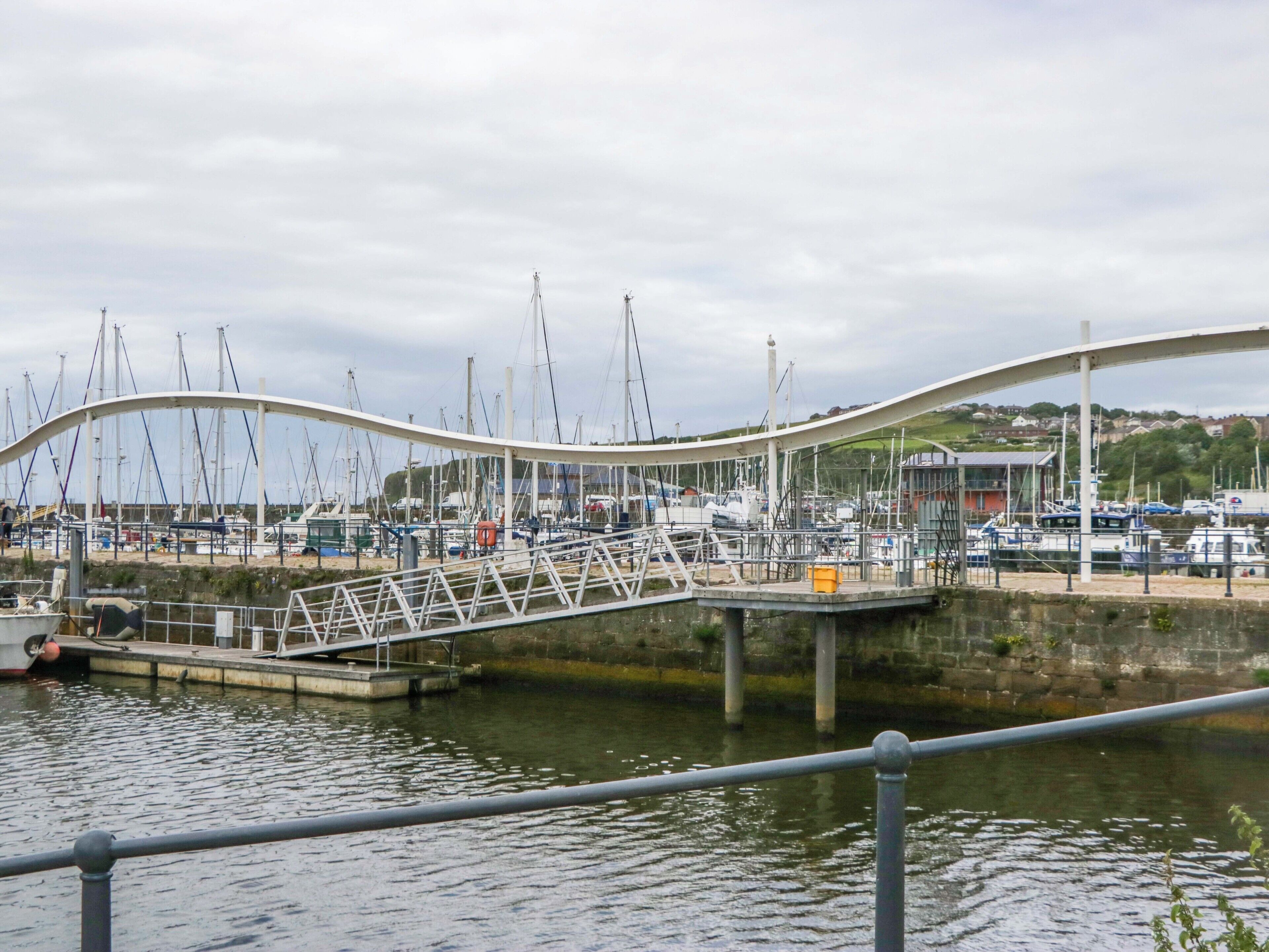Un moderno ponte sopra un porto turistico con barche ormeggiate, una passerella pedonale e un edificio sullo sfondo.