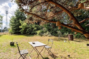 Un área de estar al aire libre con una mesa y sillas, rodeada de árboles y un gran árbol con faroles colgantes.