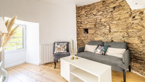 A living room with a stone wall, a grey sofa, a white coffee table, and a chair with a floral pillow. - Holiday Home 'Le Moulin De Pen Hoêt' with Shared Garden (Vertou)