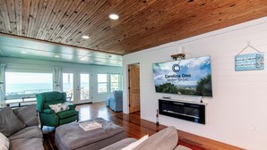 A spacious living room with a wooden ceiling, a large flat-screen TV, and a view of the ocean.
