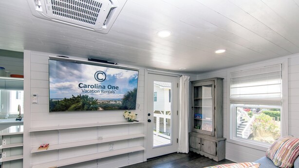 Amplia habitación con una televisión grande montada en la pared que muestra 'Carolina One Vacation Rentals'. Hay un banco de madera con cojines, una mesita y una ventana con vistas al exterior.