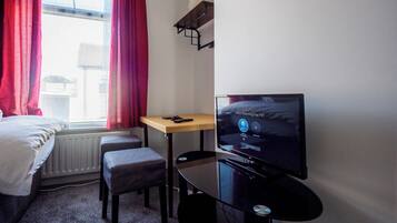 A small hotel room with a bed, a desk, a TV, and a window with red curtains.