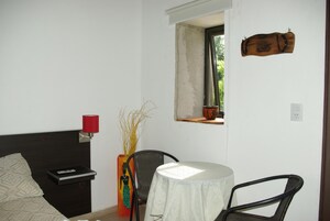 Un dormitorio con cama, mesita de noche con control remoto, una silla, una pequeña mesa con mantel blanco, una lámpara roja y ventana con vistas al verdor.