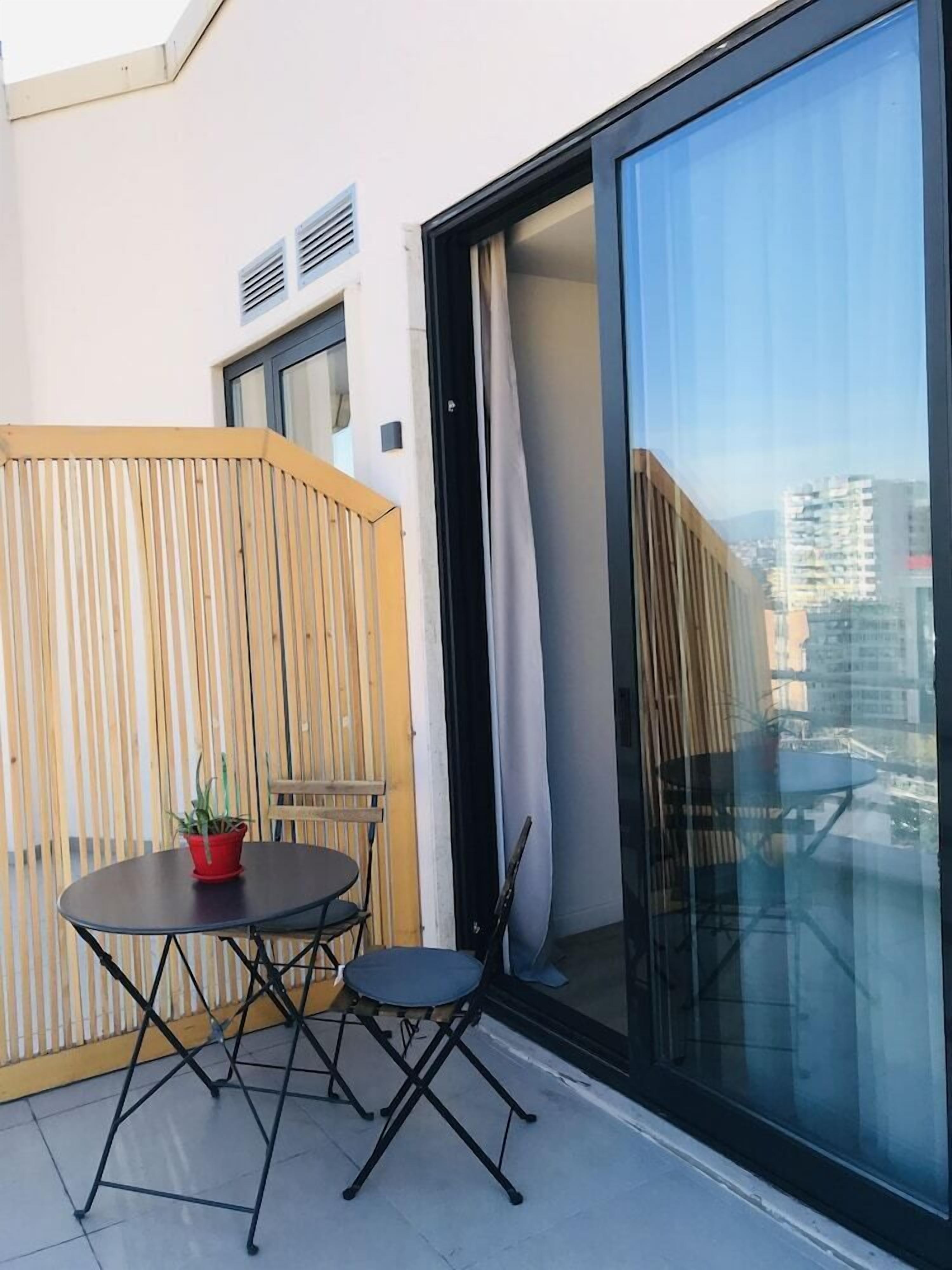 A balcony with a table and chairs, a potted plant, and a view of the cityscape through large glass doors.