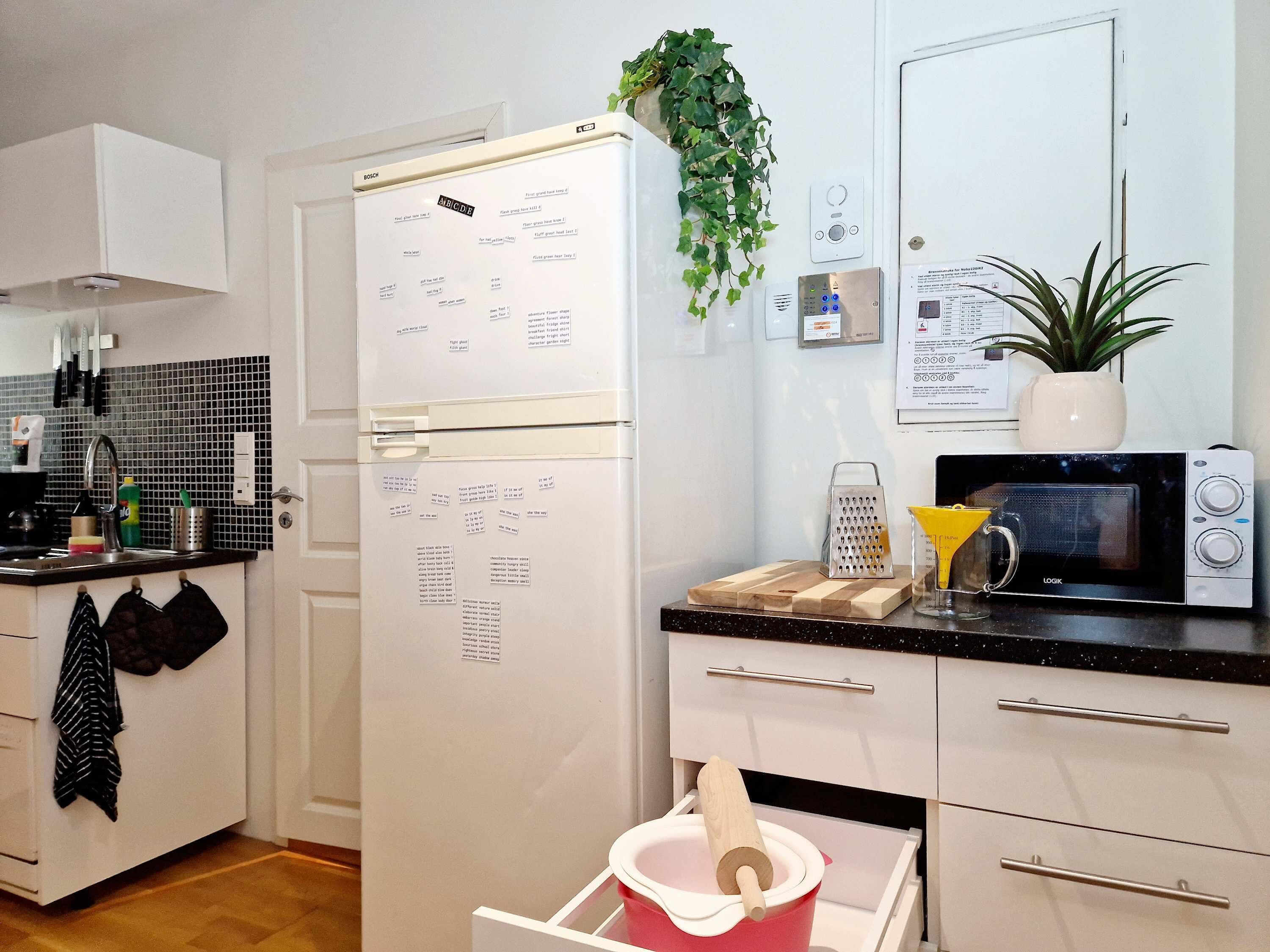 A modern kitchen with a white refrigerator, microwave, and a wooden cutting board. There is a plant on the wall and a set of knives on the counter.