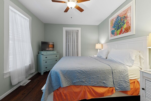 Une chambre à coucher avec un lit, un ventilateur de plafond, une télévision, une commode et un tableau au mur.