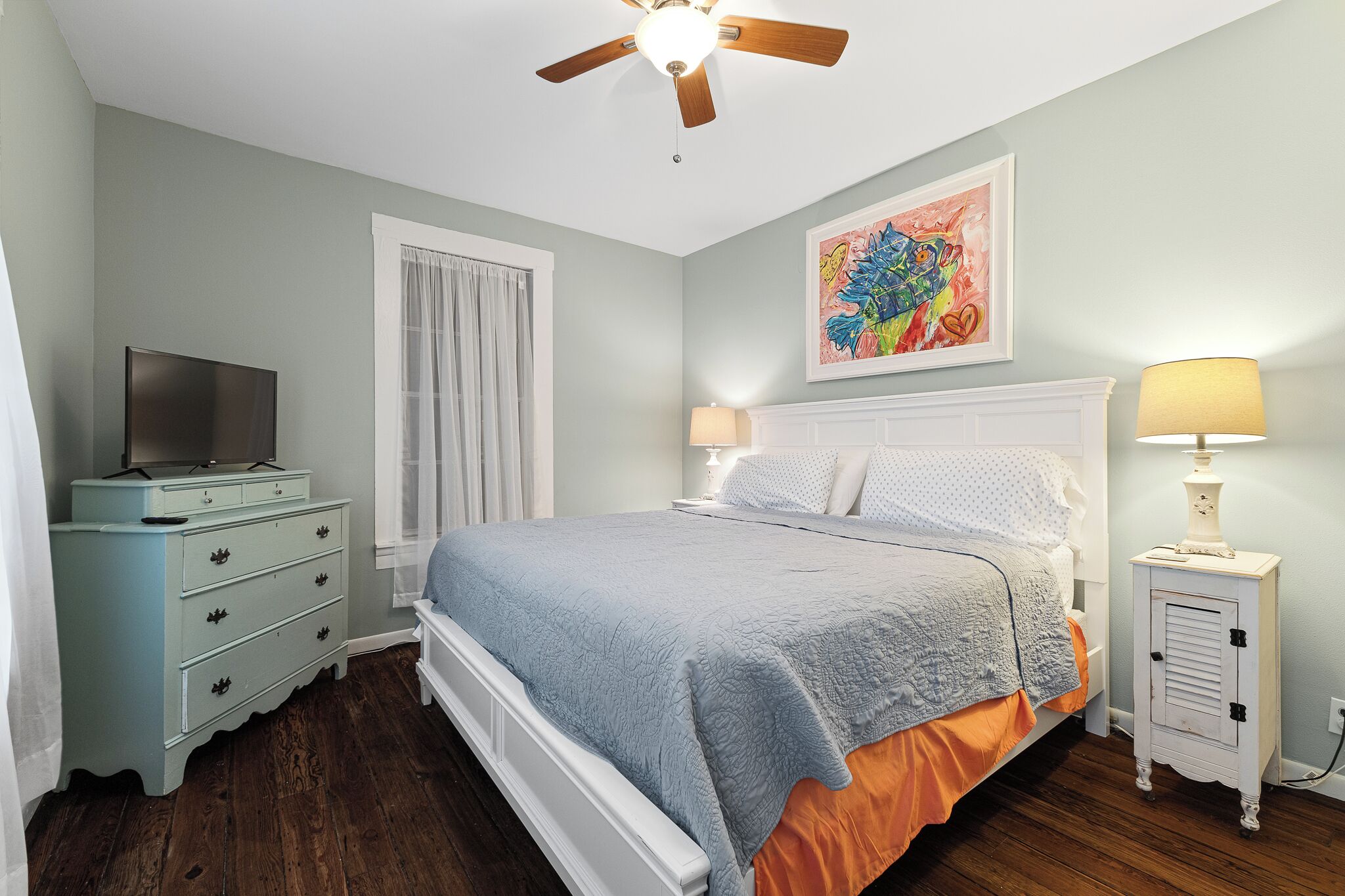 A bedroom with a bed, a dresser, a television, a ceiling fan, and a painting on the wall.
