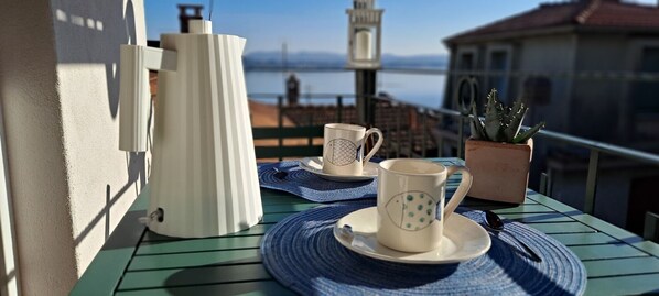 A table with a coffee pot, two cups, and a potted plant on a balcony overlooking a body of water and a bridge.