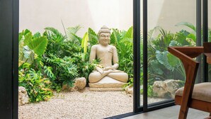 Eine Statue einer sitzenden Figur, umgeben von üppigem Grün und Steinen, sichtbar durch ein Fenster.