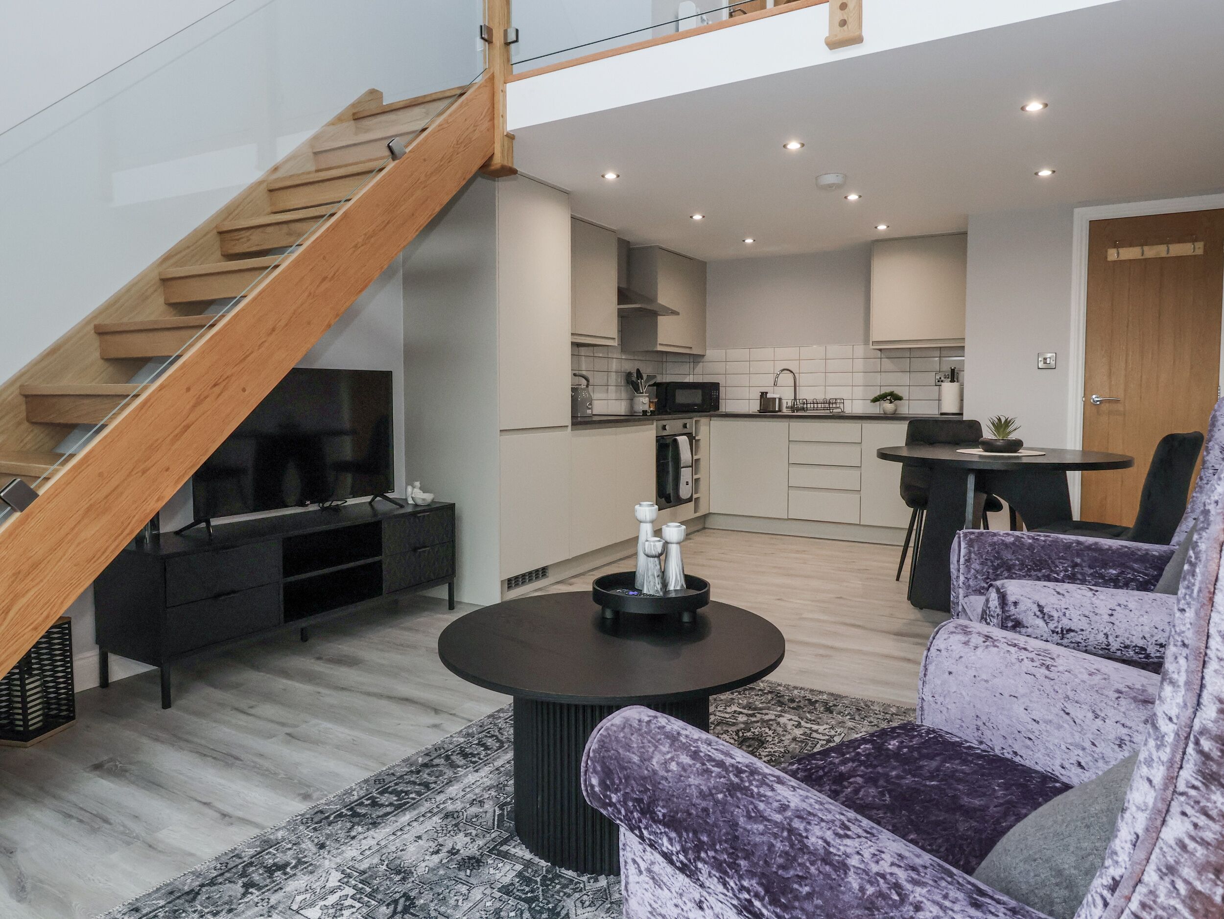 Un apartamento moderno con una escalera de madera, una sala de estar con sofá y mesa de centro, una cocina con armarios blancos, y un comedor con mesa y sillas.