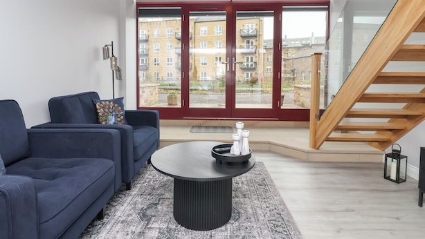 Un salon moderne avec un canapé bleu foncé, une table basse ronde et un tapis à motifs. Une porte vitrée donne accès à un balcon d’où l’on peut admirer la vue sur les immeubles.