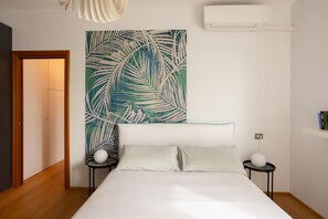 A bedroom with a bed, a wall art of palm leaves, a white air conditioner, and a small table with a spherical object.