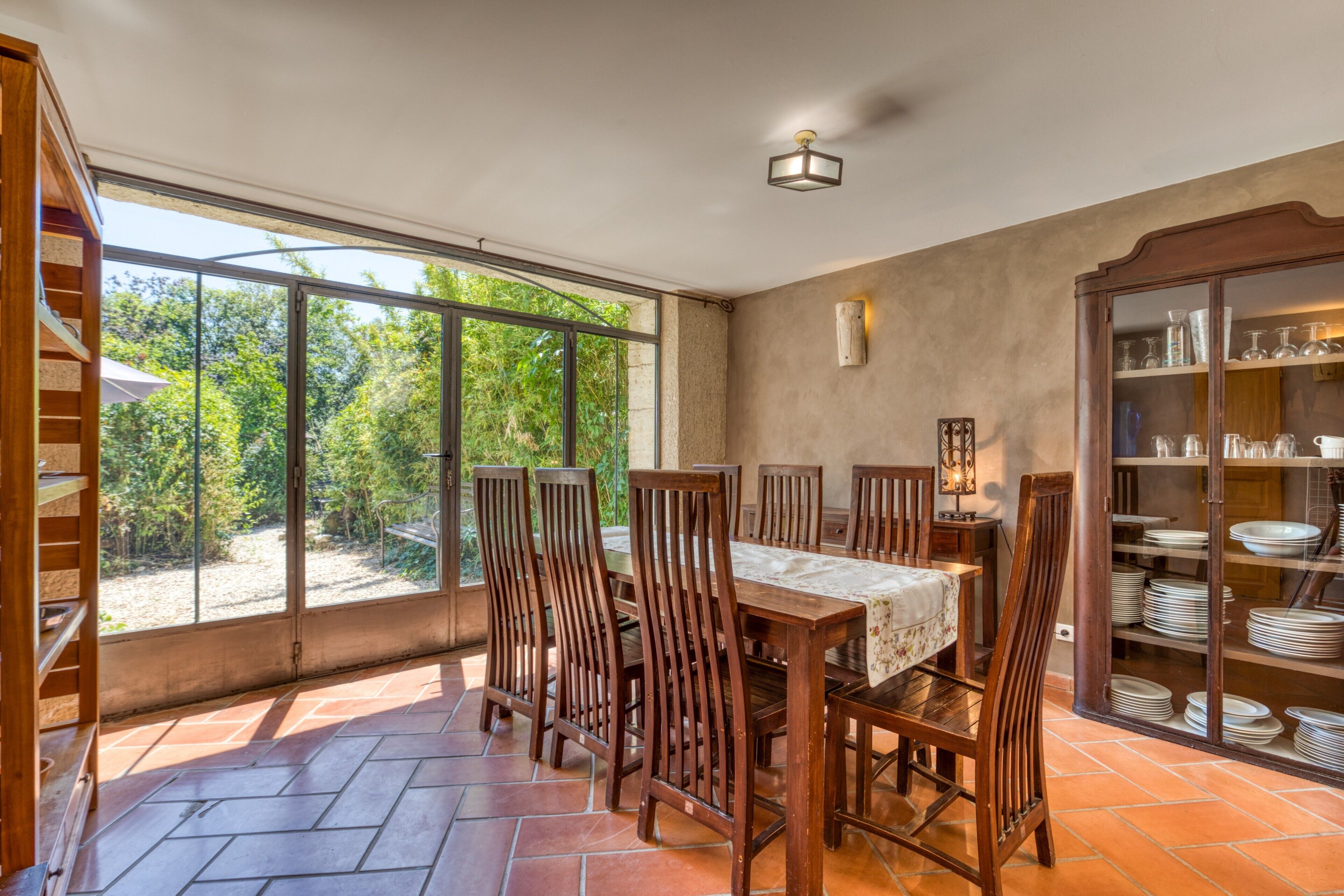 A dining area with a table and chairs, a glass cabinet with dishes, and a view of greenery through large windows.
