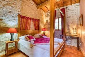 A rustic bedroom with a wooden bed, a nightstand, a desk, and a stone wall.