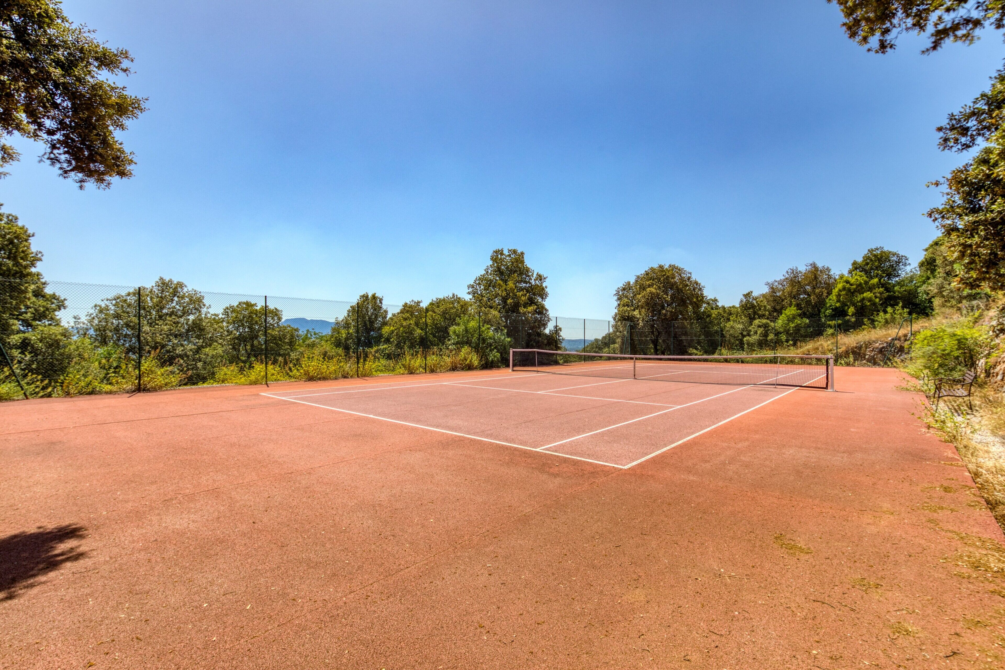 En tennisplan med nät, omgiven av träd och en klar himmel.