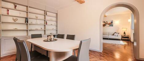 A dining area with a white table and chairs, a wooden floor, and a bookshelf with various items.