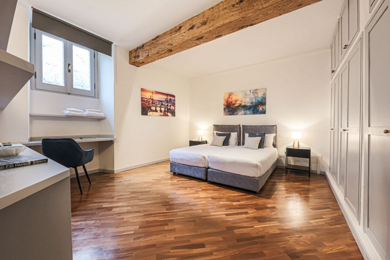 A bedroom with a wooden ceiling, a bed with pillows, a desk with a chair, and artwork on the walls.