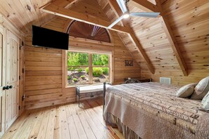 Ein Schlafzimmer in einer Holzhütte mit einem Bett, einem an der Wand montierten Fernseher und einem Fenster mit Blick auf Grünflächen.