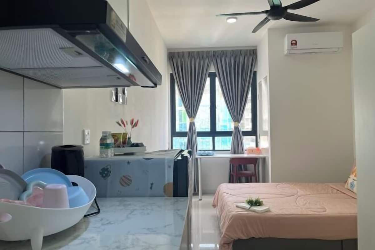 Une chambre d’hôtel compacte comprenant un coin cuisine, un lit avec une couvre-lit rose, un ventilateur de plafond et une fenêtre avec des rideaux.