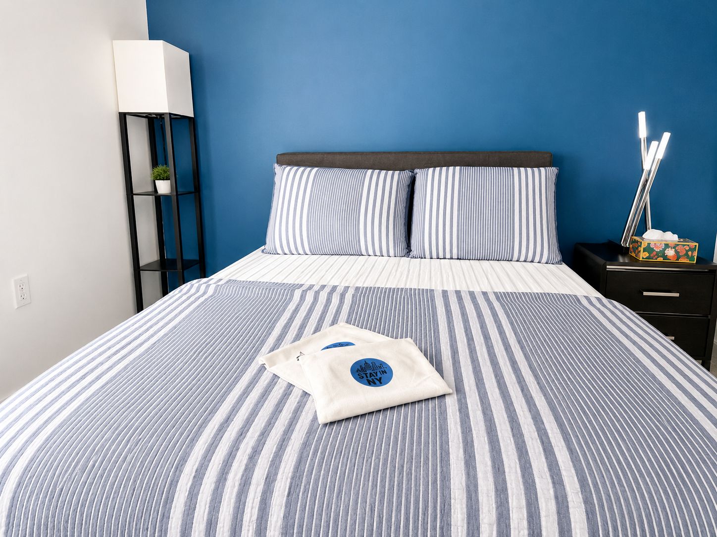 A bed with striped bedding, a pillow with a logo, a bedside table with a lamp, and a nightstand with a tissue box.