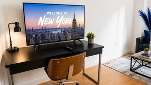 Een moderne kamer met een bureau, een stoel, een televisie waarop 'WELCOME TO NEW YORK' staat en uitzicht op de stad.
