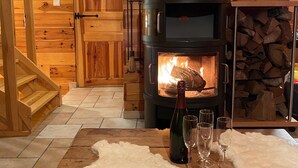 Interior de una cabaña de madera con estufa de leña, una mesa de comedor con copas de vino y una escalera.
