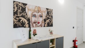 Une pièce avec une œuvre d’art accrochée au mur, une console en bois sur laquelle sont posés un vase et une bouteille, un extincteur et une porte.