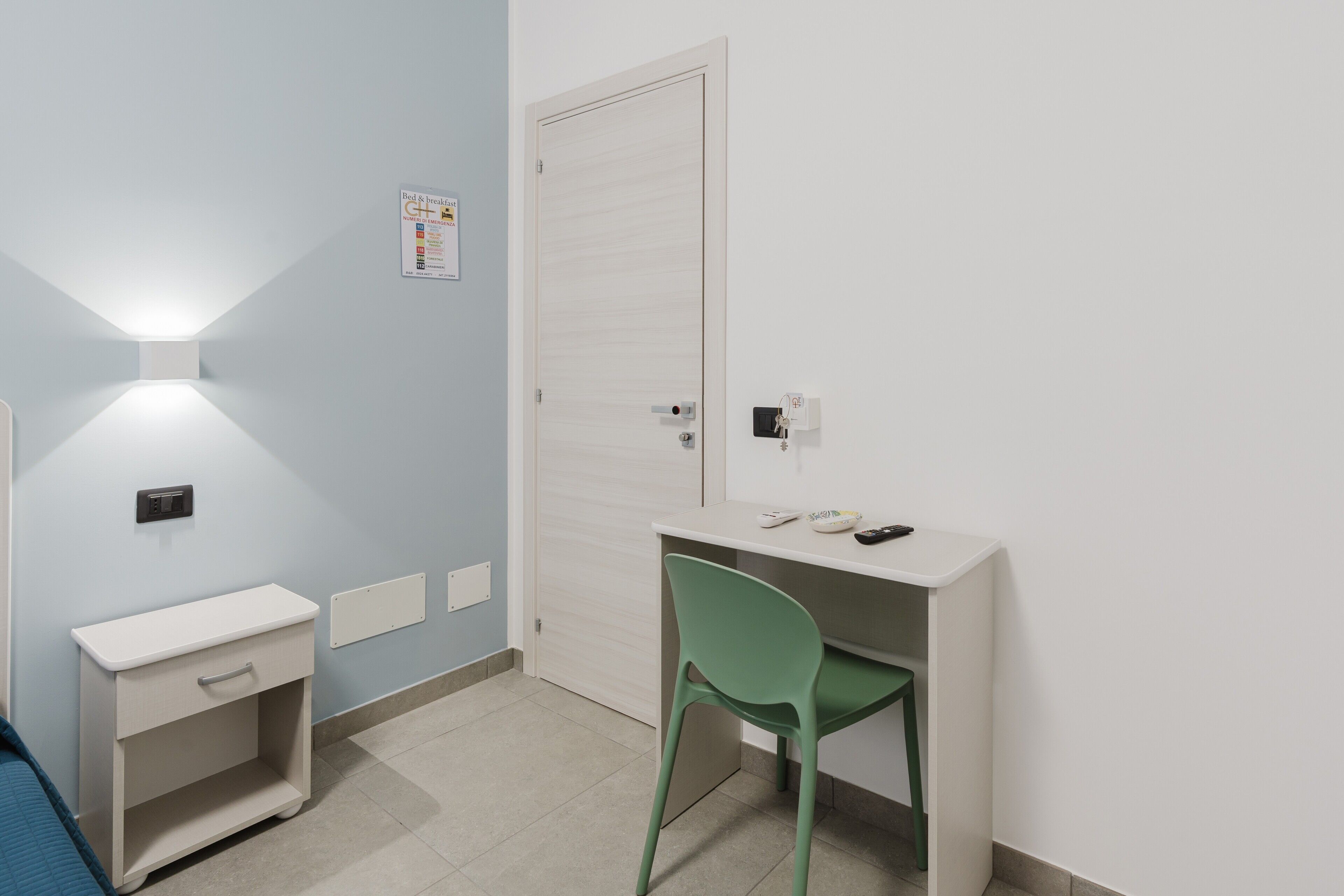 Habitació amb escriptori, cadira, llit, llum penjada a la paret, porta i presa elèctrica.