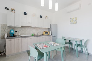 Cocina compacta con armarios blancos, microondas y cafetera. Hay dos mesas con sillas y un refrigerador pequeño.