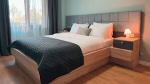 Ein modernes Schlafzimmer mit einem großen Bett, einem Nachttisch mit Lampe und Blick auf Bäume durch das Fenster.