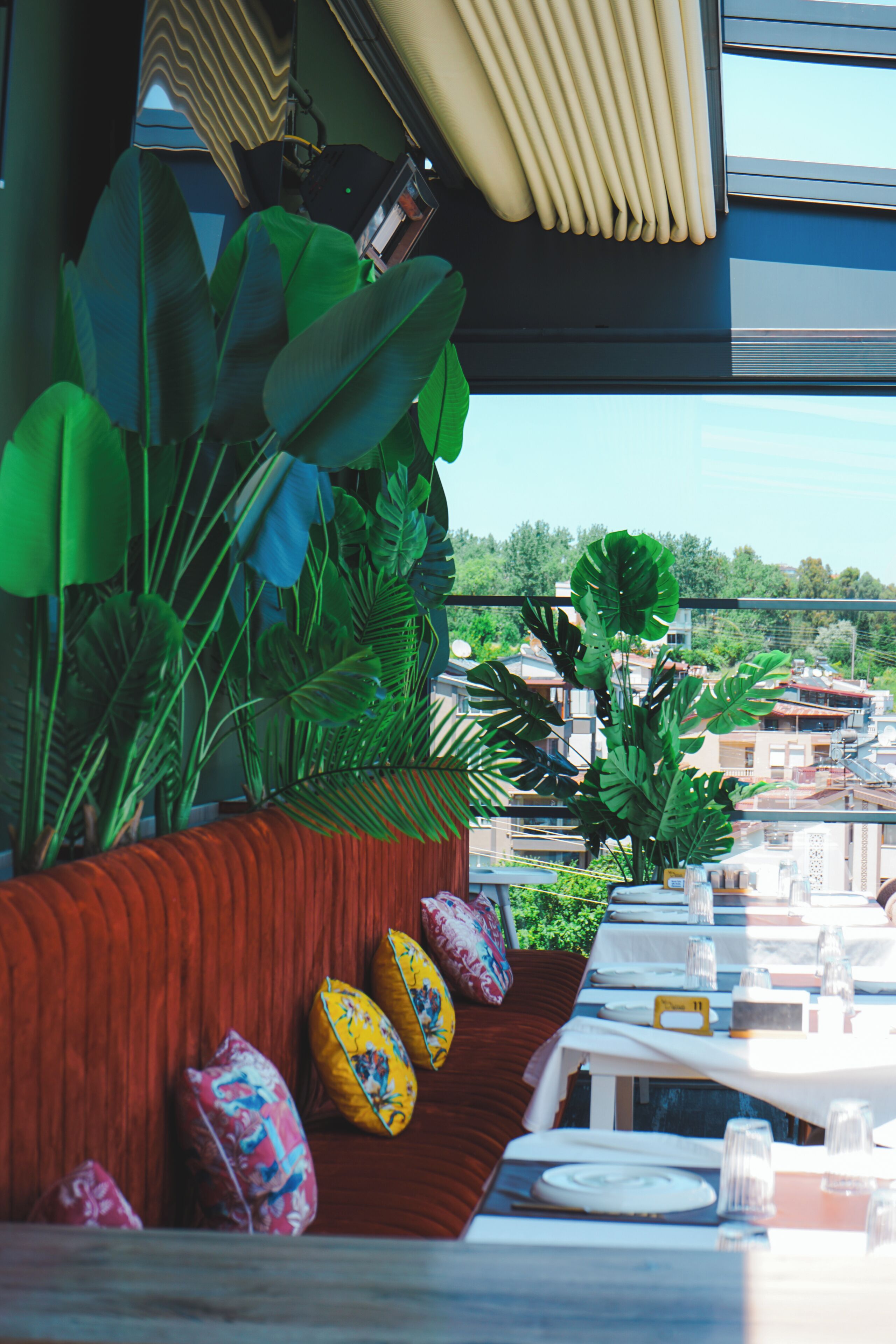 Un restaurante con una larga mesa, plantas verdes y vistas a edificios.