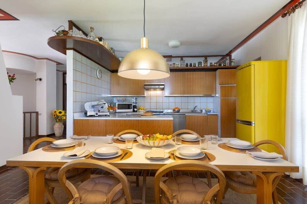 A dining area with a wooden table set for six, a yellow refrigerator, and a kitchen in the background.