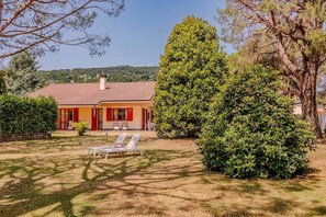 Ein Haus mit rotem Dach, ein Garten mit einem großen Baum und zwei weißen Liegestühlen.
