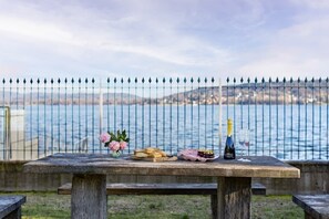 A stone table with food and drinks by a waterfront, with a metal fence in the background. - Lesa Villas (Lesa)