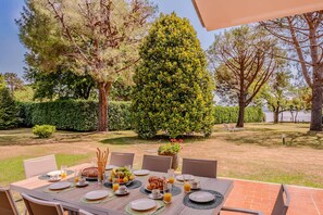 An outdoor dining area with a table set for breakfast, surrounded by trees and a hedge. - Lesa Villas (Lesa)