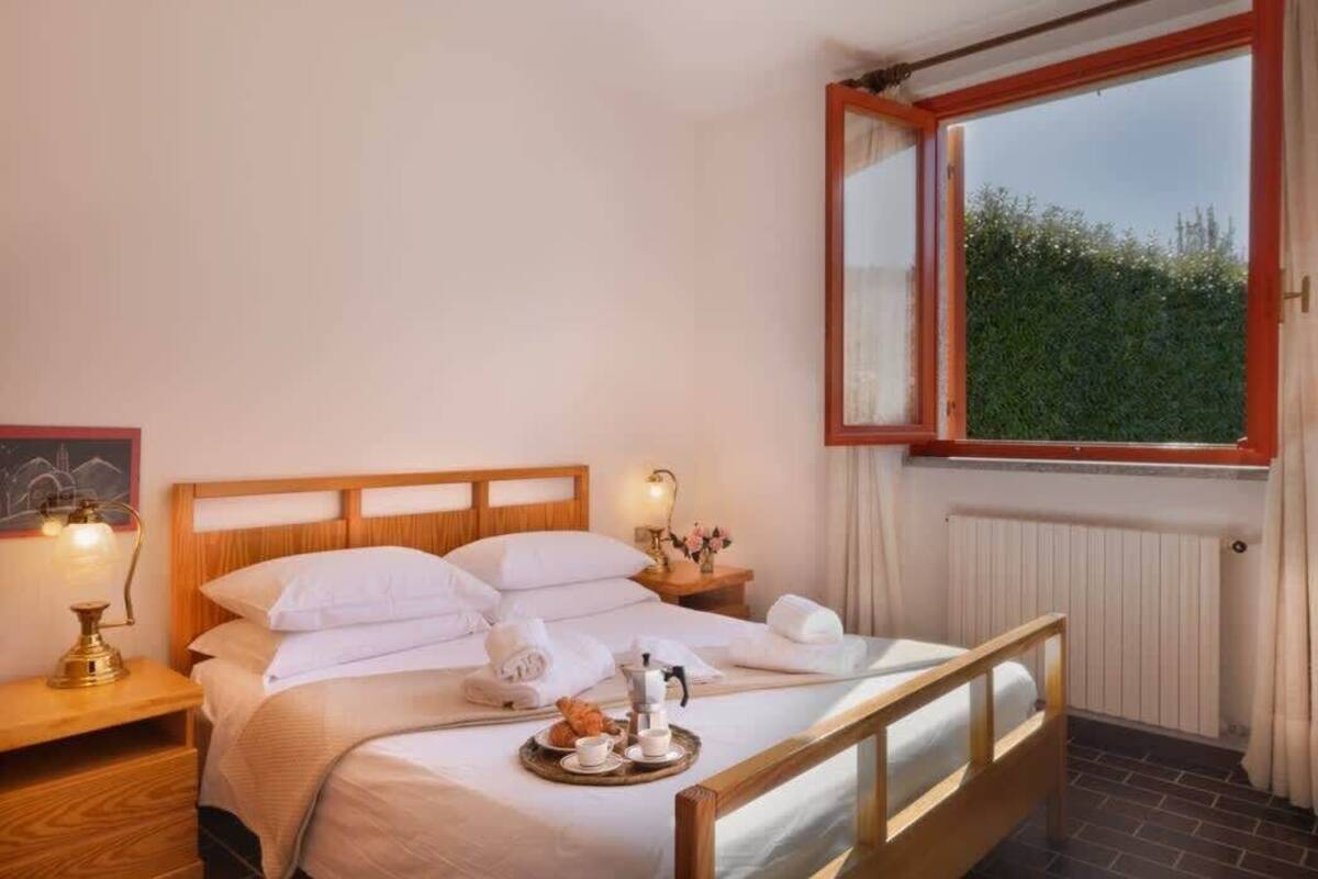 A neatly made bed with white linens, a tray with croissants and a coffee pot, and a nightstand with a lamp.