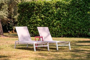Två vita loungestolar med röda ben, placerade på en gräsyta med en tät, grön häck i bakgrunden.