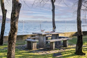 A picnic table with benches in a park by a lake, surrounded by trees and a metal fence. - Lesa Villas (Lesa)