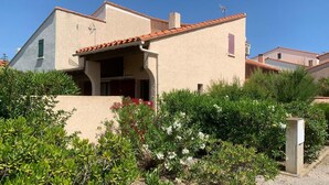 Une maison à deux étages, avec une façade de couleur beige, un toit en tuiles et un petit balcon. Un jardin, où l’on trouve diverses plantes et fleurs, se trouve devant la maison.