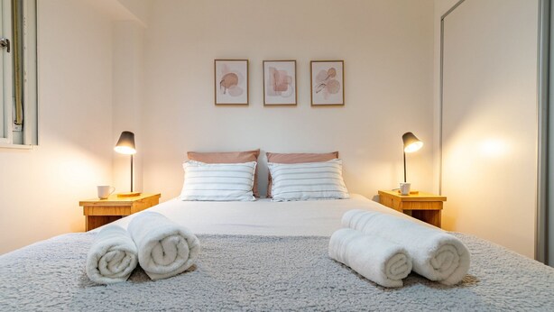 Een netjes opgemaakt bed met witte lakens en twee opgerolde handdoeken. Twee nachtkastjes met lampen, en drie ingelijste kunstwerken aan de wand boven het bed.