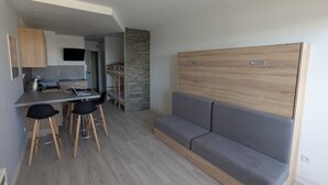 Un espacio habitable moderno y compacto con kitchenette, un sofá cama y un comedor con taburetes.