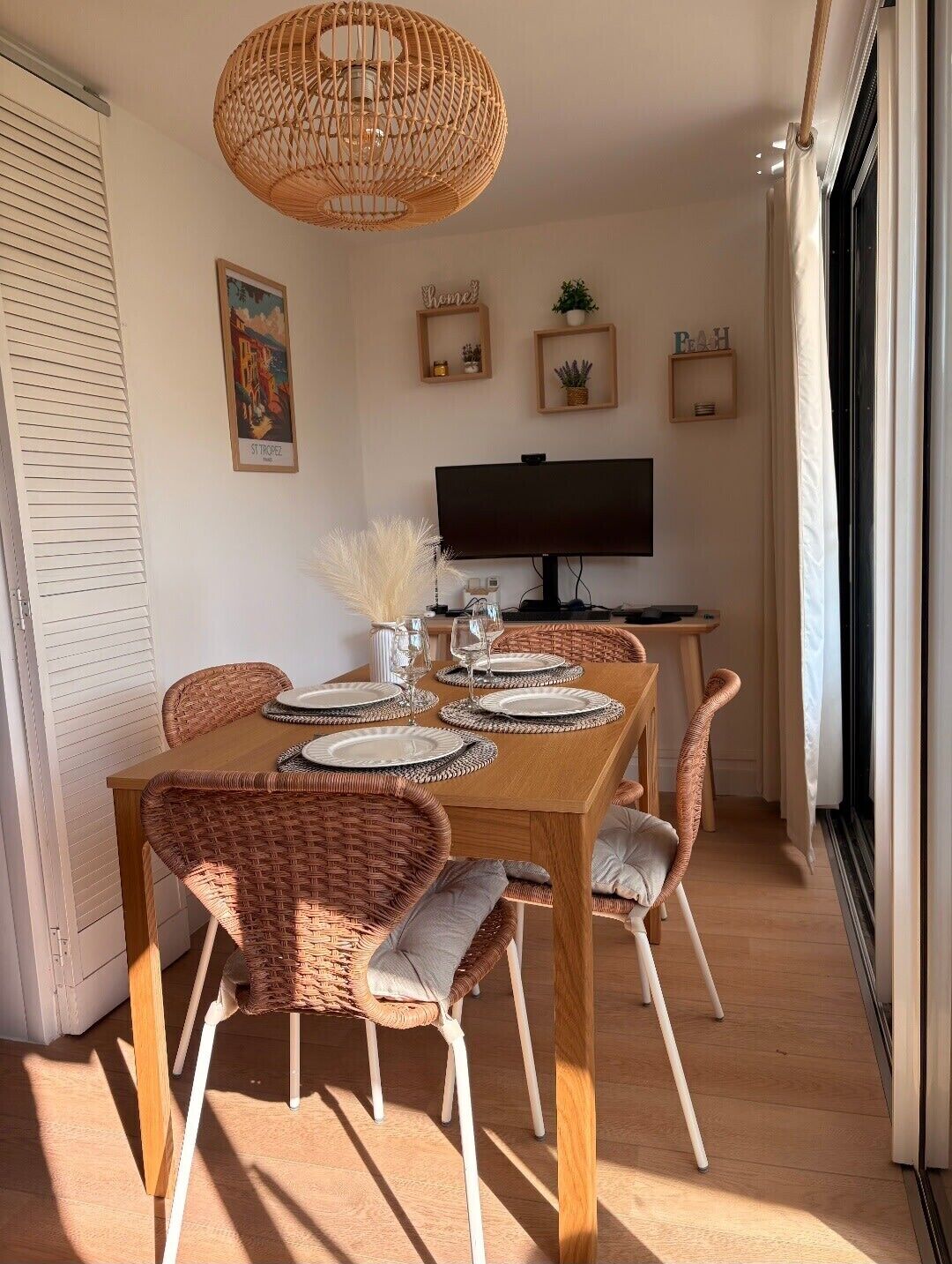 Ein Essbereich mit einem Holztisch, gedeckt für vier Personen, umgeben von Korbstühlen mit Kissen. Eine gewebte Leuchte hängt von der Decke, und ein Flachbildfernseher ist an der Wand befestigt.