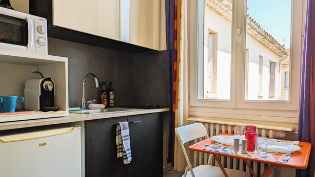 Eine kompakte Küche mit Mikrowelle, Kaffeemaschine und Spüle. Ein kleiner Tisch mit Stühlen und ein Fenster mit Blick auf Gebäude.