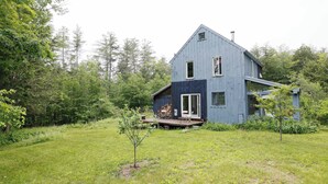 Una casa azul de dos pisos con un balcón, rodeada por un jardín cubierto de césped y árboles.