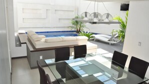A modern indoor pool area with a glass table and chairs, a white sofa, and lounge chairs. - Hotel Teocalli Tula (Tula de Allende)